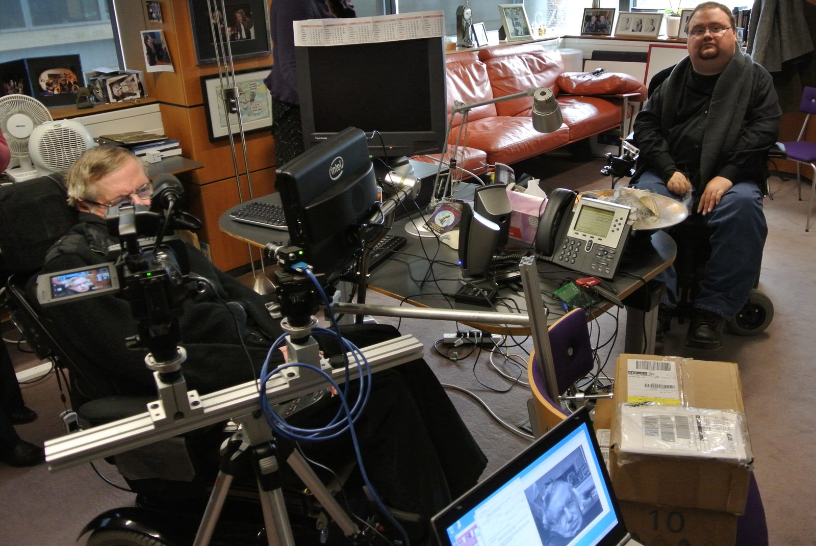 Stephen Hawking's office in Cambridge during a field testing visit. Hawking sits in his wheelchair at left, facing a camera rig and an Intel branded monitor mounted on his chair, and Pete Denman sits in his power wheelchair across the room.