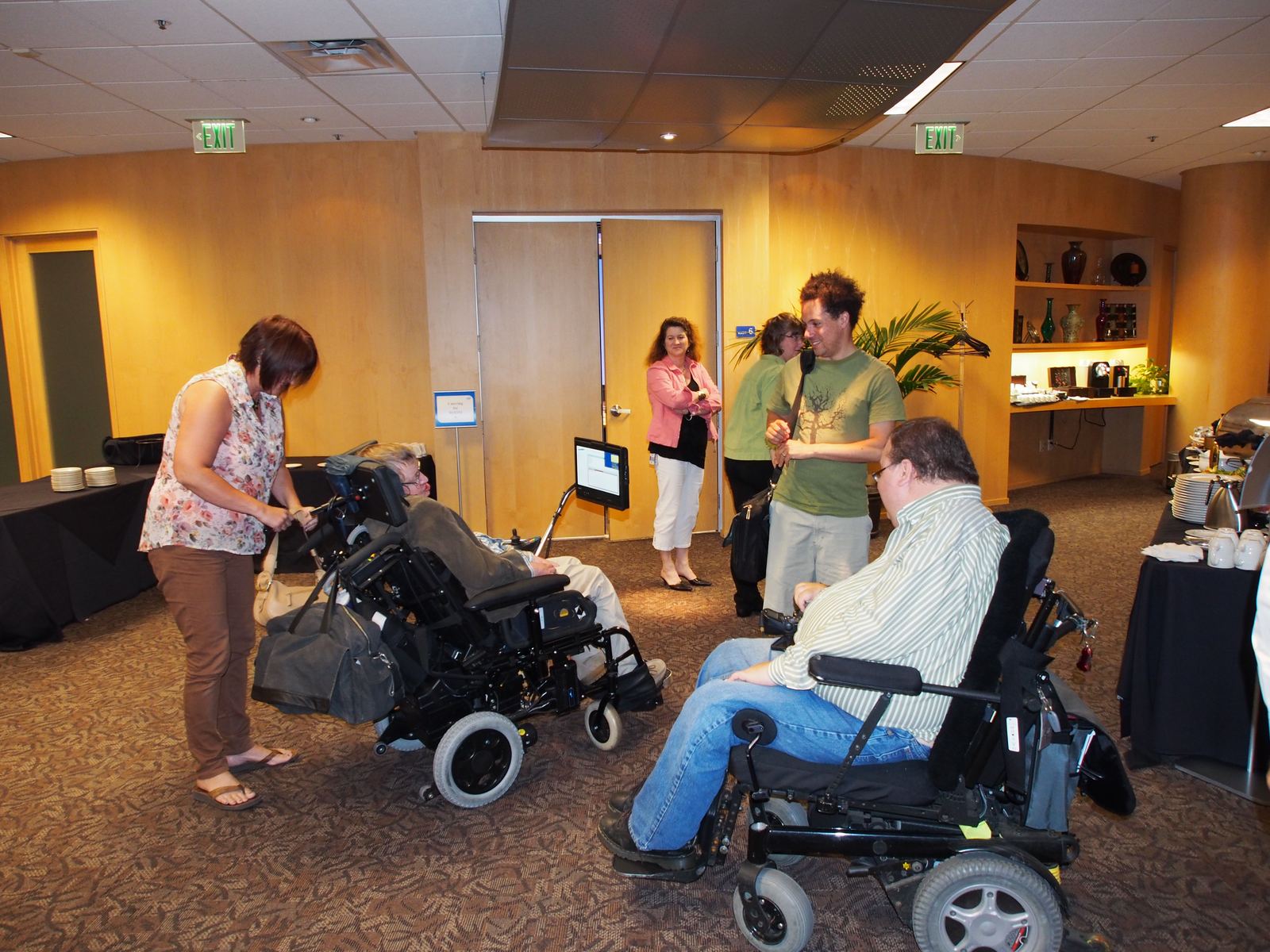 Pete Denman and Stephen Hawking side by side in their wheelchairs in a meeting room while team members prepare equipment around them.