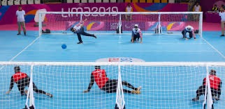 A goalball match in progress at Lima 2019: three defenders in red lie across the width of the near goal while a player in blue on the far side prepares to throw.