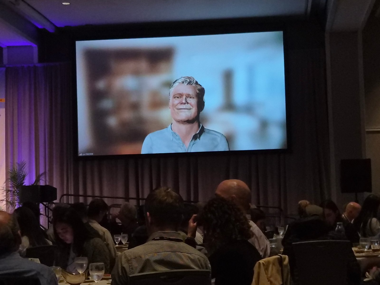 A hotel ballroom full of seated attendees looks up at a large screen showing Lex's animated avatar, a smiling man with gray hair in a light blue shirt, against a softly blurred room.