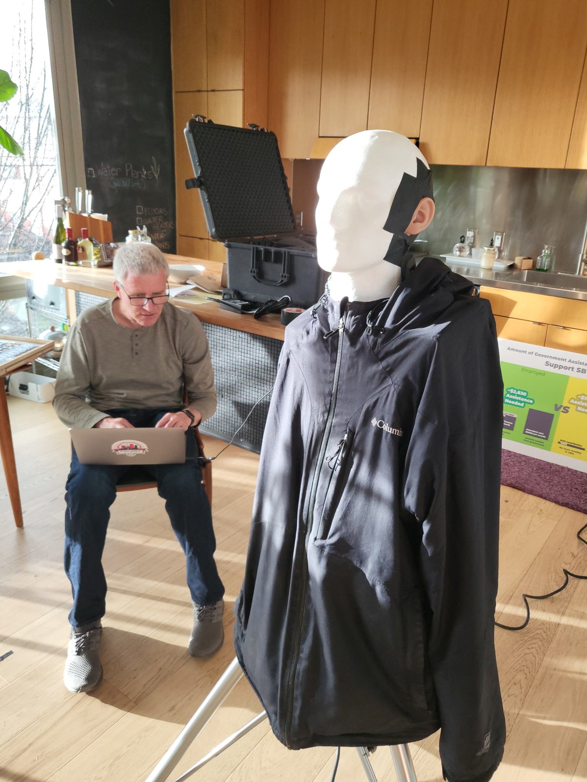 Marco Beltman sits with a laptop on his knees in a sunlit kitchen, running audio calibration, while a foam stand-in head on a tripod, dressed in a jacket with taped-on microphone ears, faces the room.