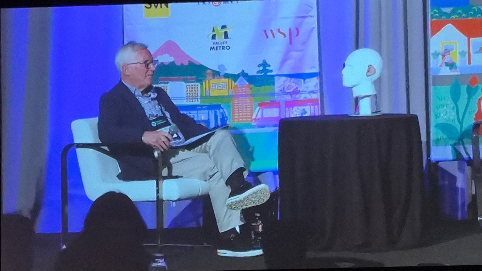 On a conference stage, a seated interviewer turns toward the Virtual Lex robot head, a white head on a small draped table, which faces him back. Sponsor logos and a colorful transit mural fill the backdrop.