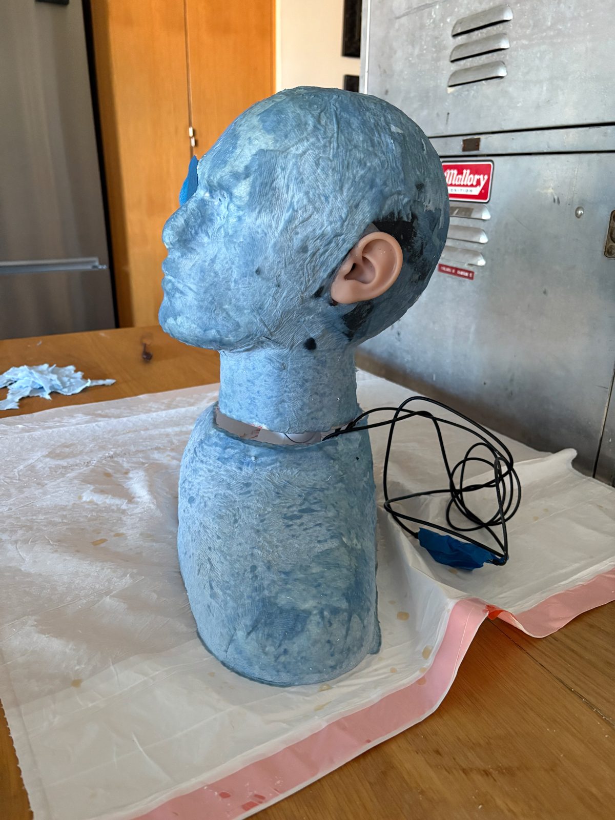 Side view of the Virtual Lex prototype bust on a worktable, showing the realistic silicone ear that houses a binaural microphone, with its wiring coiled behind the shoulder.